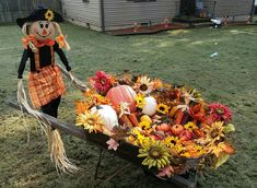 a scarecrow is pulling a wheelbarrow filled with pumpkins and flowers on the lawn