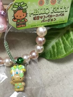 a hello kitty necklace with pearls and a green bow on it's head is sitting next to a leaf