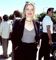 a woman standing in front of a crowd wearing black pants and a gray top with her hands in her pockets