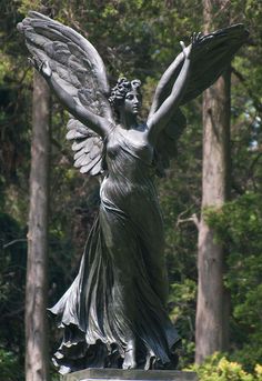 a statue of an angel with outstretched wings in front of some trees and bushes on a sunny day