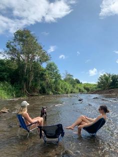 two women sitting in chairs on the edge of a river with their dogs looking at them