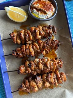 several skewers of meat and some dipping sauce on a blue tray next to lemon wedges