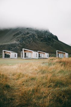 three small white buildings sitting on top of a grass covered field next to a mountain