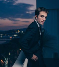 a man in a suit standing on a balcony overlooking the city at night with his hands on his hips