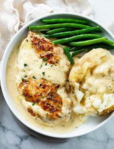 a white bowl filled with chicken and mashed potatoes covered in gravy next to green beans