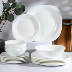 white dishes are stacked on top of each other in front of a vase with green leaves