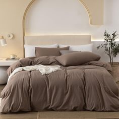 a bed with brown sheets and pillows in a room next to a white table on which there is a potted plant