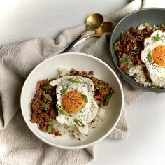 two bowls filled with meat and eggs on top of a white cloth next to spoons