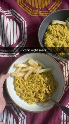 two bowls filled with noodles and french fries on top of a red table cloth next to a hand holding a fork