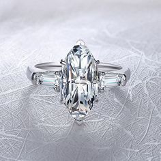 Captivating tapered baguette gem are bar-set in this elegant ring, brilliantly framing the center marquise cut moissanite. With a delicate band, this ring evokes classic beauty. Features Moissanite was originally found in meteorites(Chemical name: Silicon Carbide). It was first discovered in 1893, while a scientist was examining meteor samples from a crater in Arizona. After many years, the experts has been recreated moissanite in the laboratory, that make the gemstone with friendly, and has ete Marquise With Baguette Side Stones, Three Stone Marquise Cut Moissanite Ring, Silver Three Stone Marquise Diamond Ring, White Marquise Cut Three Stone Ring, Silver Marquise Cut Diamond Ring With Baguettes, Tapered Baguette Engagement Ring, Baguette Side Stones, Sapphire Side Stones, Baguette Engagement Ring