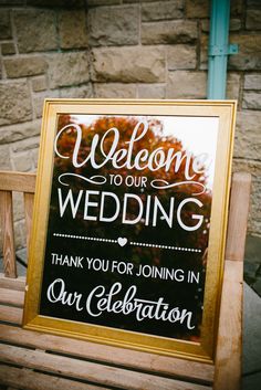 a wedding sign on a bench in front of a brick wall with trees behind it