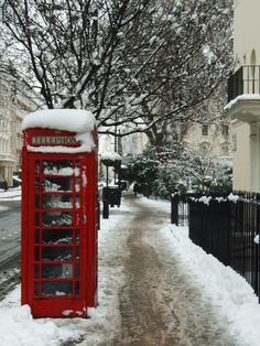 a red phone booth covered in snow next to a tree and fence on a snowy street