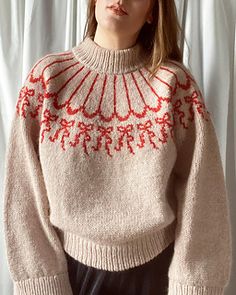 a woman standing in front of a white curtain wearing a beige sweater with red trim