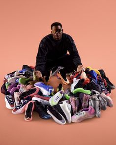 a man sitting on top of a pile of shoes in front of a pink background