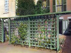 a green fence with plants growing on it in front of a building and another building