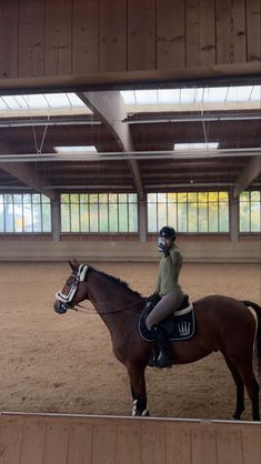 a woman riding on the back of a brown horse in an indoor arena with lots of windows