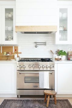 a stove top oven sitting inside of a kitchen next to a wooden stool and white cabinets