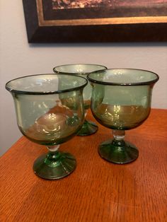 three green glass goblets sitting on top of a wooden table next to a painting