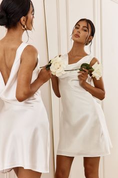 a woman in a white dress is looking at her reflection in the mirror and holding a flower