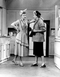 two women standing in a kitchen talking to each other and one is wearing a dress