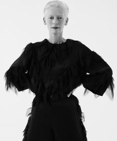 an older woman with white hair wearing a black dress and feathers on her shoulders, standing in front of a white background