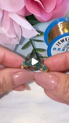 a person holding a ring in their hand with pink flowers behind it and a bottle of jewelry on the other side