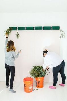 two women standing next to each other with plants in buckets on the wall behind them