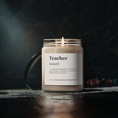 a candle sitting on top of a wooden table next to a black mug with a teacher written on it