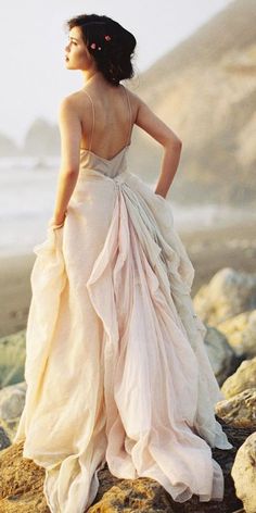 a woman in a wedding dress standing on rocks near the ocean with her back to the camera