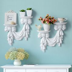 two white vases with flowers are on the wall next to a dresser and shelf