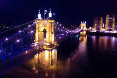 a bridge that is lit up at night with lights on it and buildings in the background