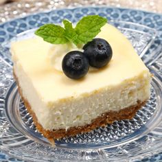 a piece of cheesecake on a glass plate with two blueberries and mint sprigs