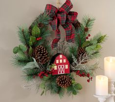 a wreath with pine cones, evergreen leaves and a red barn ornament is hung on the wall