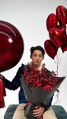 a man sitting on a couch holding a bunch of red balloons and a bouquet of flowers