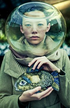 a woman holding a fish in her hands and wearing a large glass bowl over her head