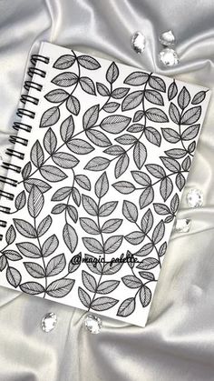 a spiral notebook sitting on top of a white satin covered bed sheet with leaves drawn on it