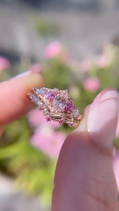 a hand holding a ring with pink and blue stones on it in front of some flowers