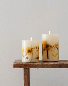 two white candles with yellow flowers on them sitting on a wooden table next to a wall