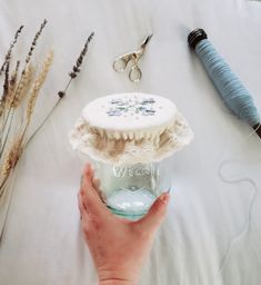 a person holding a jar with some thread and scissors next to it on a bed