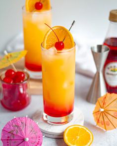 two glasses filled with orange juice and garnished with cherries next to cocktail umbrellas