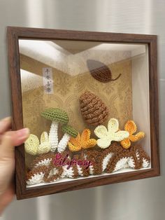 a person holding up a small wooden frame with crocheted flowers and pine cones