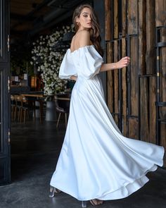 a woman in a white dress is standing near a wooden wall and looking off to the side