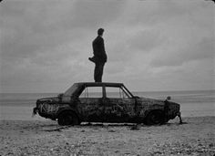 a man standing on top of an old car in the sand at the beach with graffiti all over it