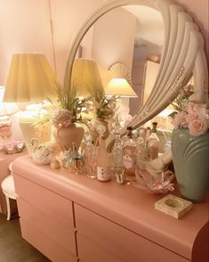 a pink dresser topped with lots of bottles and vases filled with flowers next to a mirror