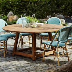 a wooden table with blue chairs around it on a brick patio surrounded by greenery