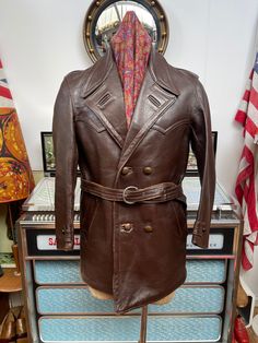 "Here we have a Vintage double breasted belted leather coat. Now I am not 100%sure if it's date and origin,have been selling Vintage since 2001 and my firs thought was 1950s but I am not completely sure,stitching detail,lapels and moleskin lining tells me 1950s but the belt looks later and there is a date on the inside of left front pocket,1978,I just think it's that late,the pocket and main lining is original but the sleeves and inner pocket have been replaced ( See pics). Nicely worn in leather in places,patina looks really nice,wear to leather around normal places,shoulders,hem,arms and top of the back as pictured,really thick coat,very warm and heavy. Definitely made for the colder weather,perfect on a vintage motorcycle. Fits a small to 40\" maybe 41\" with just a t-shirt underneath. Vintage Long Leather Jacket For Business, Brown Trenchcoat, Fitted Vintage Outerwear With Belt Loops, Vintage Double-breasted Leather Jacket For Fall, Fitted Vintage Belted Outerwear, Vintage Fitted Belted Outerwear, Vintage Long Brown Pea Coat, Vintage Brown Long Pea Coat, Retro Brown Double-breasted Outerwear