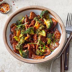 a bowl filled with meat and broccoli on top of a table next to silverware