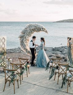 a couple standing next to each other in front of the ocean with chairs and decorations