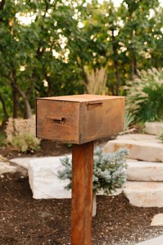 an old wooden mailbox sitting in the middle of a garden with rocks and trees