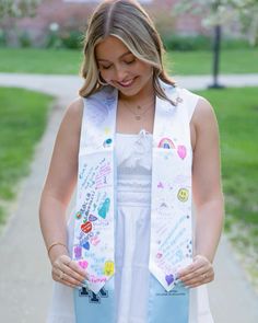 a woman wearing a white dress and a blue scarf with buttons on it is looking down at her hands